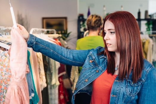 young woman with red hair, happy and enjoying a day of shopping, dressed in a blue denim jacket and a red t-shirt, choosing a pink dress from a rack, shopping in a fashion shop clothes. shopping concept. natural light from the shop window, sun rays, display with clothes, rack, shop window, clothes, Horizontal.