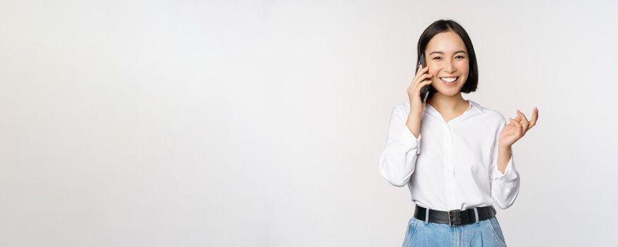 Smiling happy asian woman talking on smartphone with client, saleswoman on call, holding mobile phone and gesturing, standing over white background.