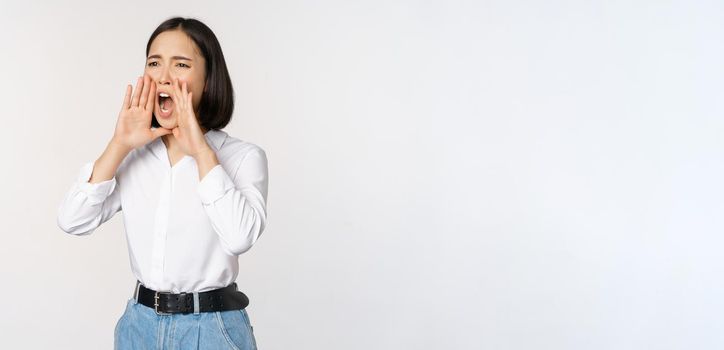 Image of young asian woman calling for someone, shouting loud and searching around, standing against white background.