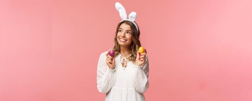 Holidays, spring and party concept. Portrait of dreamy, tender young blond girl feeling happy, celebrating religious day, holy Easter, wear rabbit ears and white dress, hold painted eggs, look away.