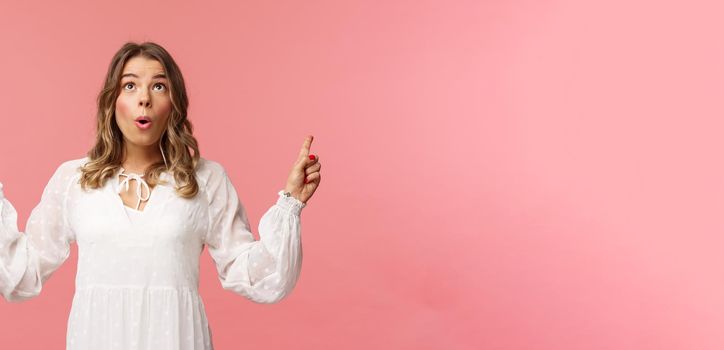 Excited beautiful caucasian blond woman in white dress, fold lips in excitement, looking pointing fingers up at something stunning and breathtaking, enjoying view, pink background.