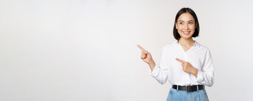 Portrait of attractive adult asian woman pointing, looking left with pleased smile, showing banner or logo aside, standing against white studio background.