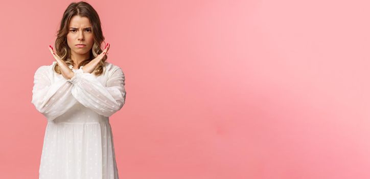 Portrait of serious-looking confident and displeased young blond woman make stop sign, cross arms over chest and frowning with disapproval, prohibit, reject bad offer, stand pink background.