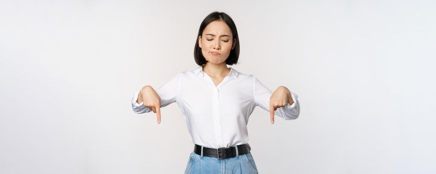 Portrait of skeptical, disappointed asian businesswoman, korean girl pointing and looking down with doubts, standing against white background.