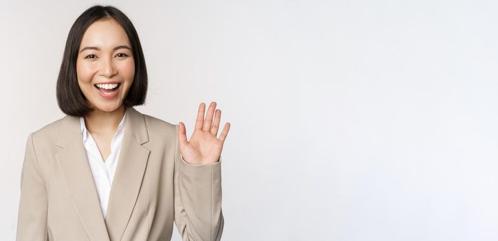 Friendly business woman, asian office lady waving hand and saying hello, hi gesture, standing over white background.