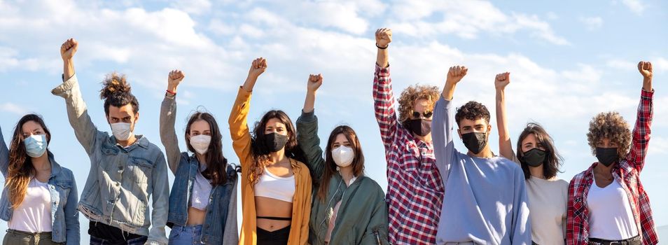 Panoramic image of multiracial activist protesters with fists raised up in the air wearing protective face mask protesting on the street. Demonstration concept.