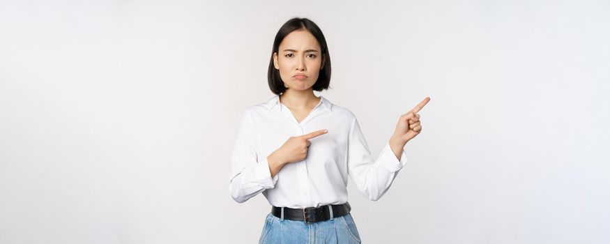 Sad and gloomy asian woman looking disappointed, complaining at banner or advertisement, pointing fingers right at promo and frowning upset, white background.