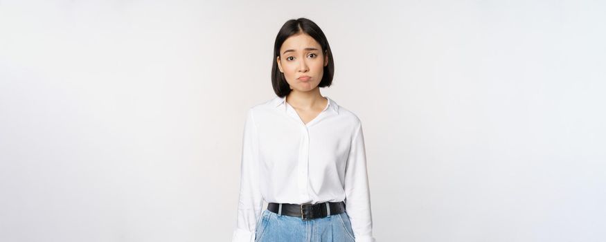 Image of sad office girl, asian woman sulking and frowning disappointed, standing upset and distressed against white background.