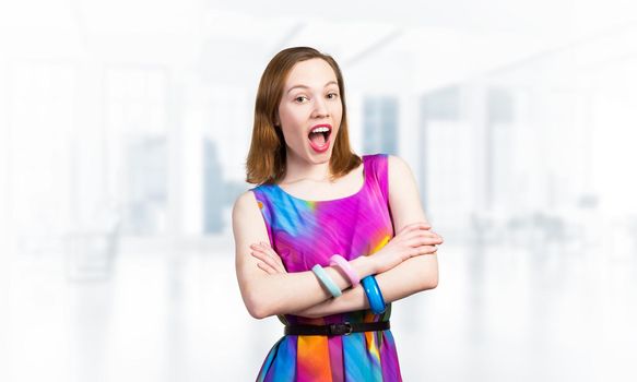 Beautiful caucasian woman standing with folded arms. Elegant fashionable lady in colorful dress and bracelets. Studio shot of good looking redhead girl with open mouth on blurred office background