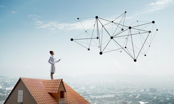 Conceptual image of young female doctor interracting with media network structure while standing on roof of brick building. Medical industry and modern technologies