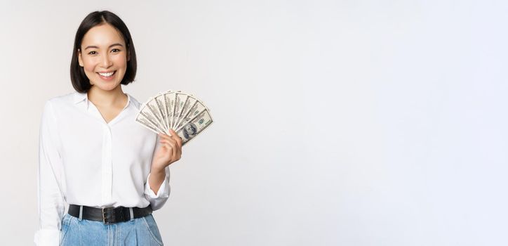 Credit and loan concept. Smiling young asian woman holding cash dollars and looking happy at camera, white background.