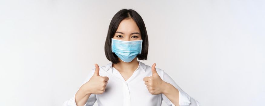 Portrait of smiling asian office lady in medical face mask, showing thumbs up, recommending smth, standing over white background.