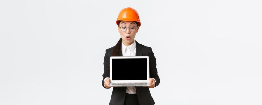 Impressed asian female construction engineer, architect or manager at enterprise showing laptop screen with amazed expression, looking at profit diagram during meeting, wear safety helmet in factory.