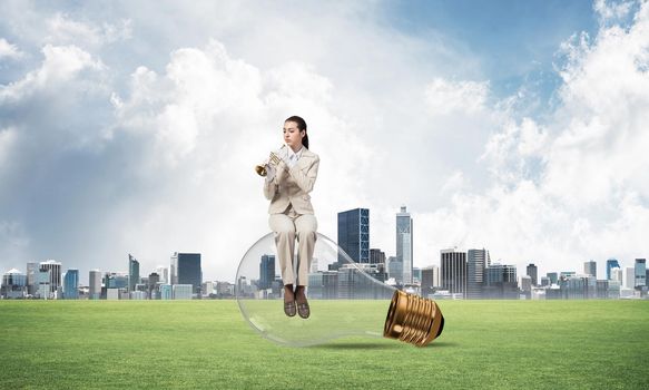 Attractive woman playing trumpet brass on green grass. Young businesslady in white suit sitting on big light bulb with music instrument on cityscape background. Musician practicing and performing