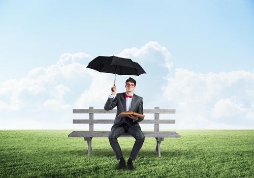 A young student holds a book and an umbrella. sits on a wooden bench