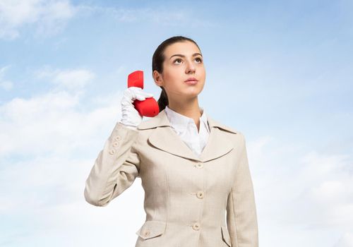 Young business woman holding red retro phone and looks upward. Call center operator in white business suit and gloves with telephone on sky background. Hotline telemarketing concept with copy space