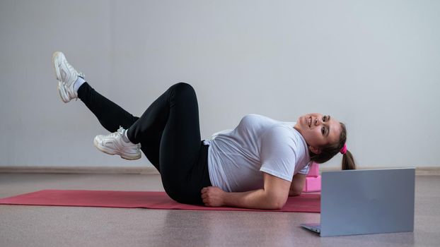 A chubby young woman is watching an online fitness lesson on a laptop. Distance sports training