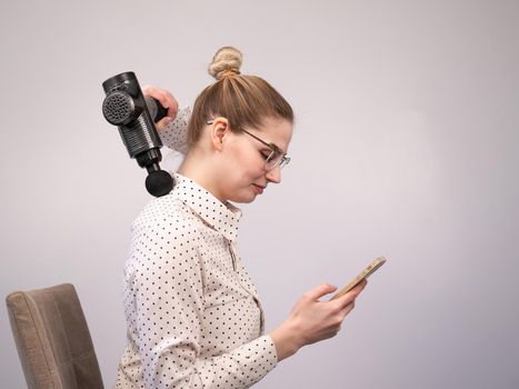 A young woman sits on a chair, uses a smartphone and makes herself massage with an electro massage gun