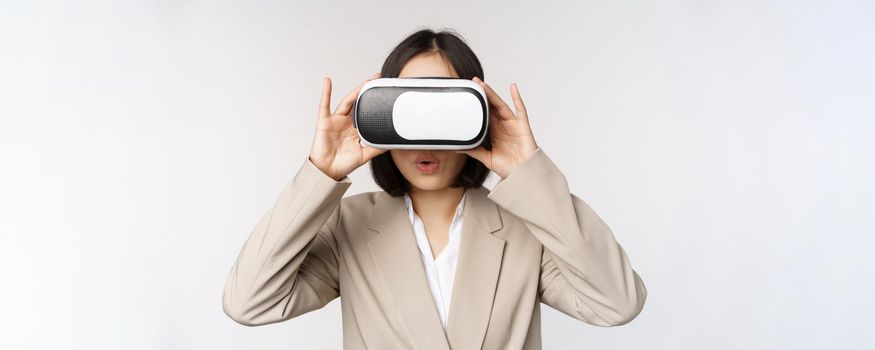 Amazed office woman, asian business person in suit, wearing vr headset, looking at smth in virtual reality glasses with impressed, wow face, white background.