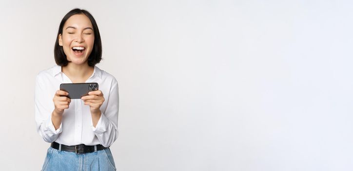 Image of young asian woman watching on smartphone app, holding mobile phone and looking at screen, laughing and smiling, standing over white background.