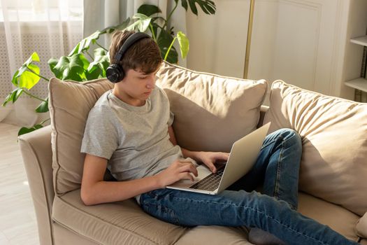 A teenager in a gray t-shirt and blue jeans, lying on a sofa in a sunny room, wearing black headphones on his head, looks into a laptop.