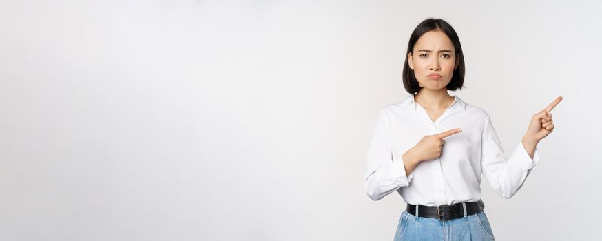 Sad and gloomy asian woman looking disappointed, complaining at banner or advertisement, pointing fingers right at promo and frowning upset, white background.