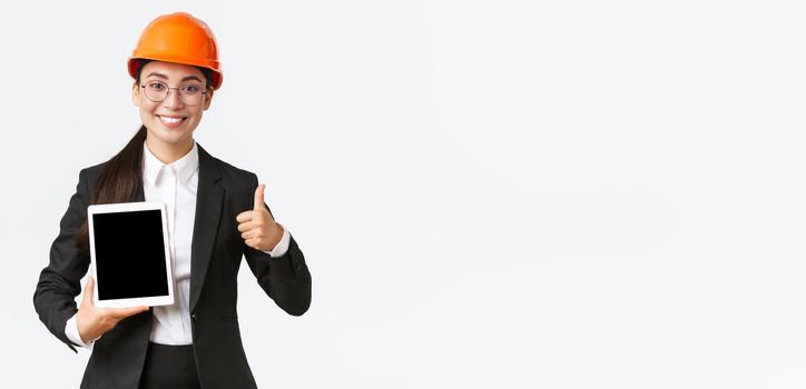 Professional smiling asian female construction manager, factory engineer in business suit and safety helmet showing thumbs-up and digital tablet screen with positive graph, white background.