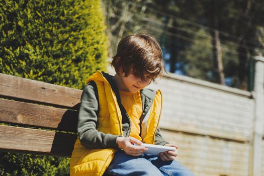 Schoolboy plays game his cell phone sitting on bench in the city park. Boy kid in yellow vest and green hoodie uses mobile gadget to chat with friends while outdoors in the garden during sunny day.