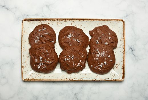 Close up several freshly baked round brownie chocolate cookies with flake salt on white porcelain plate on table, elevated top view, directly above