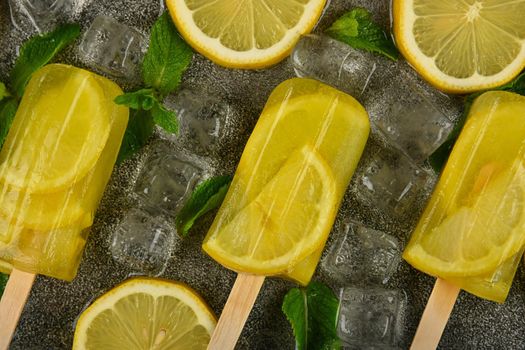 Close up frozen fruit juice popsicles with fresh lemon slices, green mint leaves and ice cubes on gray table surface, elevated top view, directly above