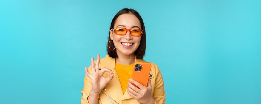 Happy korean girl in sunglasses, showing okay sign and holding mobile phone, using smartphone app, recommending application, standing over blue background.