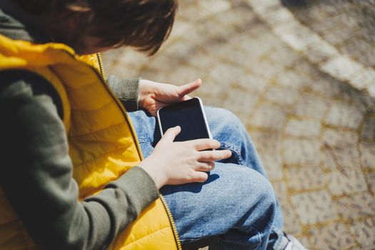 Schoolboy plays game his cell phone sitting on bench in the city park. Boy kid in yellow vest and green hoodie uses mobile gadget to chat with friends while outdoors in the garden during sunny day.