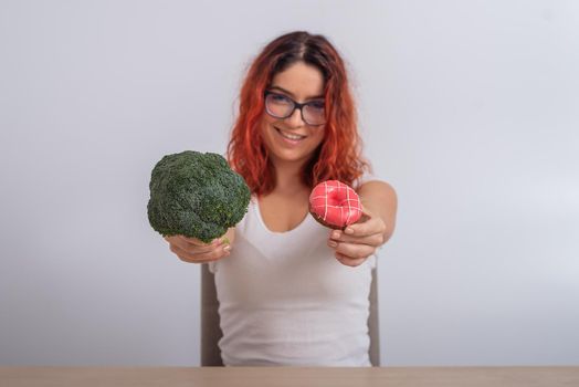 Caucasian woman chooses between vegetables and fast food. Redhead girl holding broccoli and donut on a white background