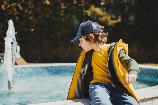 Schoolboy sitting on a fountain in public park during spring sunny day playing with water. Kid in yellow vest chilling in park with water sprinkling in the background. Summer holidays outside.