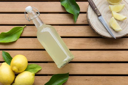 horizontal image of glass bottle full of lemonade with lemons in the lower left corner and natural cutting board with old knife and lemon slices on wooden table