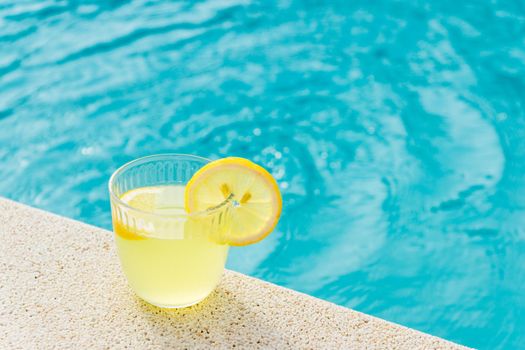lemonade in transparent glass with lemon slice on the edge of the pool with turquoise blue water background in horizontal format