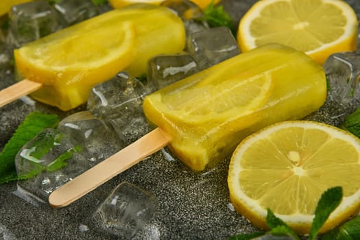 Close up frozen fruit juice popsicles with fresh lemon slices, green mint leaves and ice cubes on gray table surface, high angle view