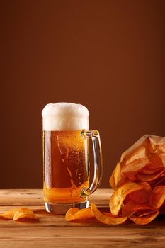 Close up one glass mug of lager beer with white froth and bubbles and paper bag of potato chips on wooden table over dark brown background with copy space, low angle side view