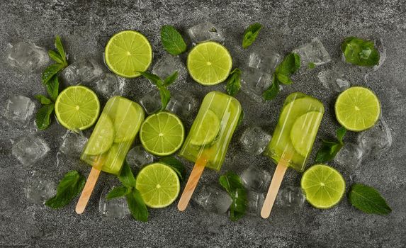 Close up several frozen fruit juice popsicles with fresh lime slices, green mint leaves and ice cubes on gray table surface, elevated top view, directly above