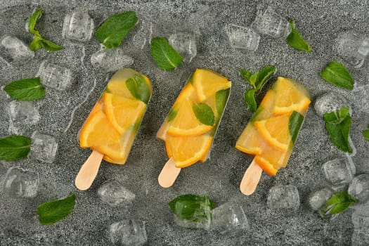 Close up three fruit ice cream popsicles with fresh orange slices, green mint leaves and ice cubes on gray table surface, elevated top view, directly above