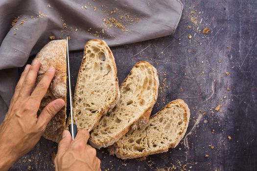 Zenithal image of an organic bread with hands with a knife breaking it into slices placed in a fan shape on a dark surface surrounded by crumbs and a gray cloth with natural light and a touch of flash