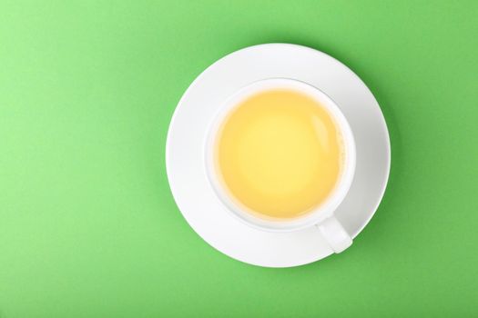 Close up one full white cup of green oolong or herbal tea on saucer over pastel green paper background, elevated top view, directly above