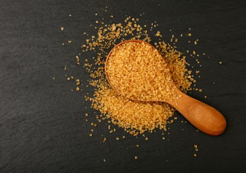 Close up one wooden scoop spoon full of brown cane sugar on black slate board, elevated top view, directly above