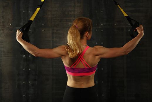 Rear view portrait of one young middle age athletic woman at crossfit training, exercising with trx suspension fitness straps over dark background