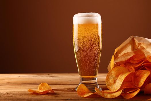 Close up one glass of lager beer with white froth and bubbles and paper bag of potato chips on wooden table over dark brown background with copy space, low angle side view