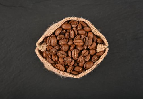 Close up one jute canvas bag of roasted Arabica coffee beans over black slate background, elevated top view, directly above