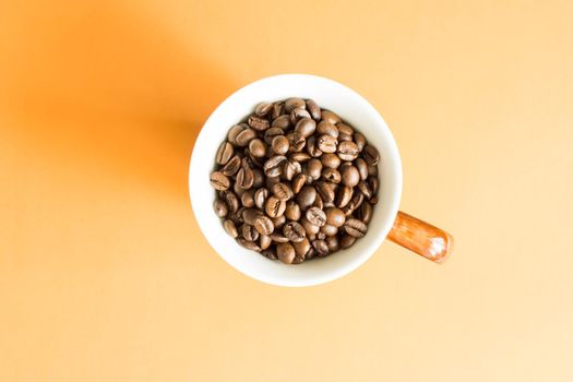 White cup with brown handle filled with coffee beans collected from above with orange background