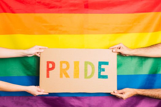 hands of man and woman holding a cardboard with the word pride written in colors on the background gay pride flag