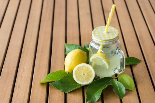 horizontal image of transparent jug with handle and straw filled with lemonade on brown wooden slat table with lemon leaves around and lemons