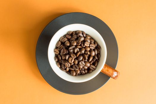 White cup with brown handle filled with coffee beans collected from above with orange background